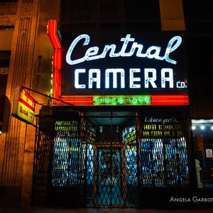 Könnte beinhalten: Ein Vintage-Ladenlokal mit einem Neon-Schild, auf dem "Central Camera Co. Since 1899" steht. Das Ladenlokal hat ein Metalltor und ein Fenster mit einem Schild, auf dem "Lanago's Must Eliminate All Cash On The Spot" steht.