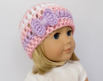 Puede incluir: Un gorro de ganchillo en tonos rosa, blanco y lavanda, adornado con un lazo lavanda. El gorro está diseñado para una muñeca. El gorro se muestra en una muñeca con cabello rubio y ojos azules, vistiendo una camiseta blanca.