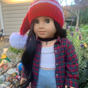 May include: A doll with long dark hair wearing a red knit hat with a white pom-pom, a red and green plaid button-down shirt over a white crop top, light blue jeans, and a black choker necklace with a silver letter S charm pendant.