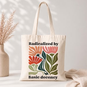 May include: A cream-colored canvas tote bag featuring the black text 'Radicalized by' at the top and 'Basic decency' at the bottom. The center design displays a colorful floral illustration with orange, red, pink, and dark green leaves and petals.