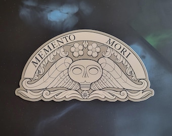 May include: A silver sticker with a skull and wings design, featuring the words "Memento Mori" in an arched banner. The sticker has a detailed, engraved appearance with floral accents, suitable for gothic or alternative themes.
