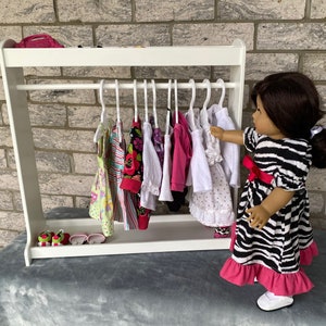 May include: A white wooden doll clothes rack with a variety of colorful dresses hanging on hangers. A doll is standing next to the rack, wearing a black and white zebra print dress with a pink ruffle.