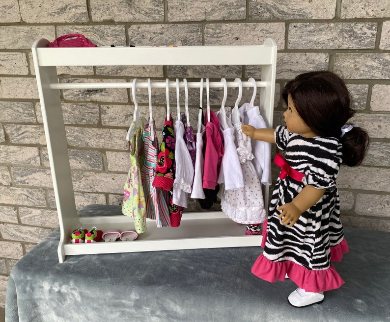 May include: A white wooden doll clothes rack with a variety of colorful dresses hanging on hangers. A doll is standing next to the rack, wearing a black and white zebra print dress with a pink ruffle.
