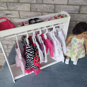 May include: A white doll clothes rack with a pink purse, shoes, and several dresses hanging on hangers. A doll wearing a green and white dress is standing next to the rack.