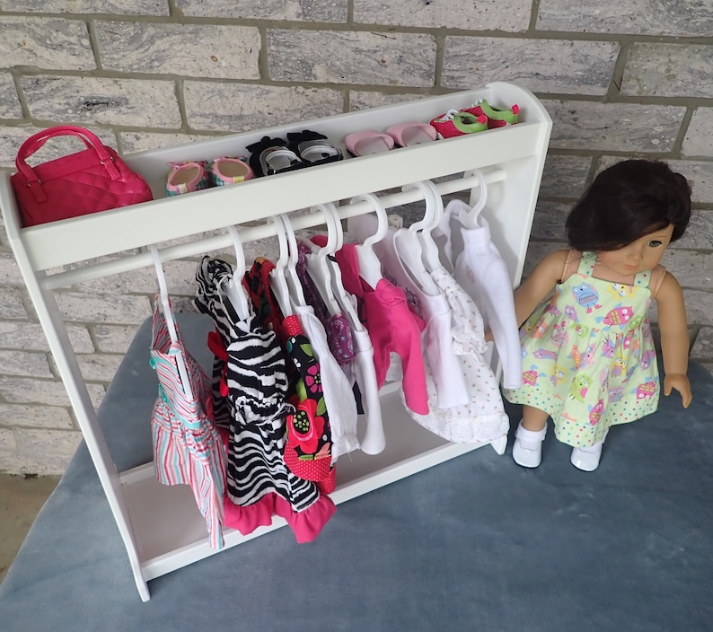 May include: A white doll clothes rack with a pink purse, shoes, and several dresses hanging on hangers. A doll wearing a green and white dress is standing next to the rack.