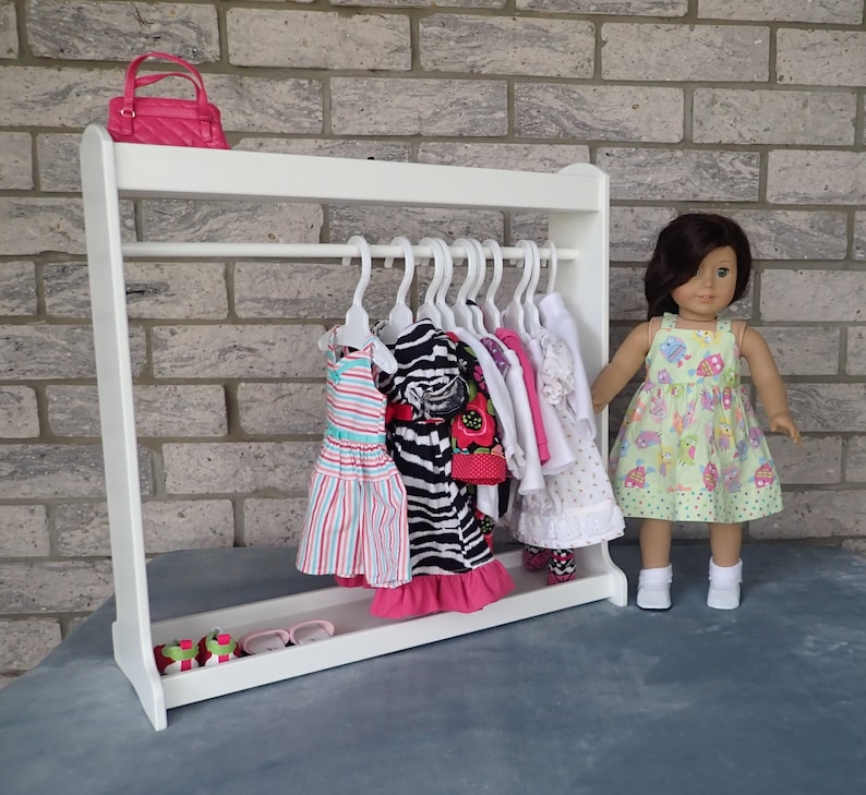 May include: White wooden doll clothes rack with a pink purse on top. The rack has several hangers with colorful dresses and a doll in a green and white dress standing in front of it.