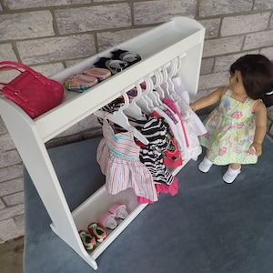 May include: White wooden doll clothes rack with a shelf on top. The rack has hangers for clothes and a shelf for shoes. There are several doll clothes hanging on the rack, including a striped dress, a zebra print dress, and a red dress with flowers. There are also several pairs of doll shoes on the shelf.