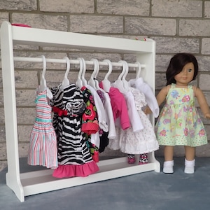 May include: White wooden doll clothes rack with a variety of colorful dresses and shirts hanging on hangers. A doll is standing next to the rack wearing a green and white dress.