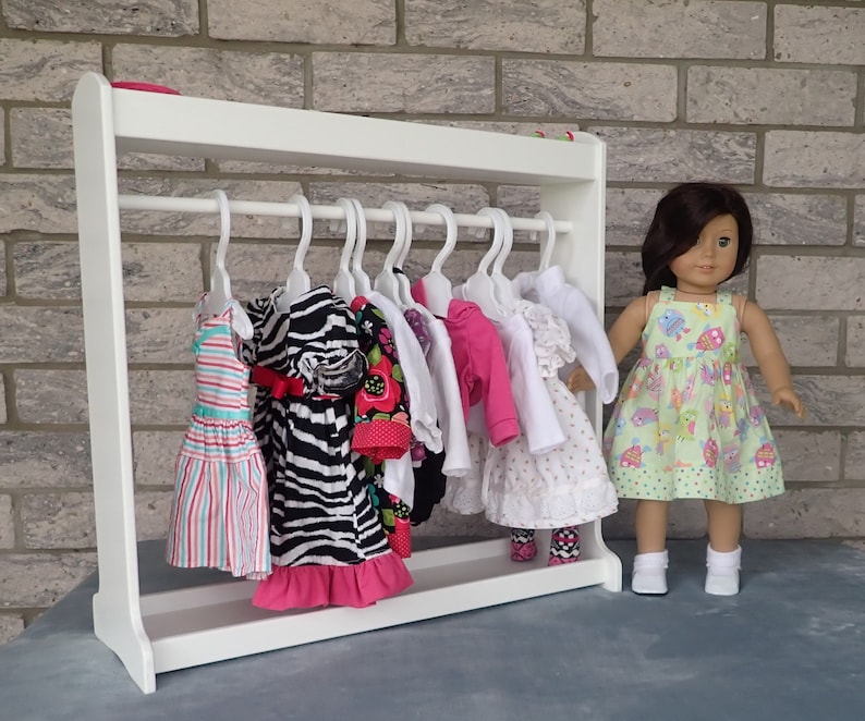 May include: White wooden doll clothes rack with a variety of colorful dresses and shirts hanging on hangers. A doll is standing next to the rack wearing a green and white dress.