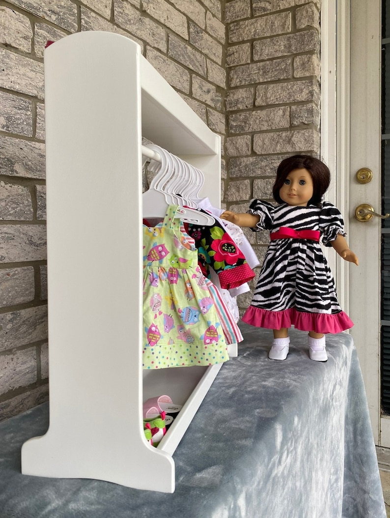 May include: White wooden doll clothes rack with a shelf on top and a hanging rod. The rack is filled with colorful doll clothes and shoes. A doll is standing next to the rack.