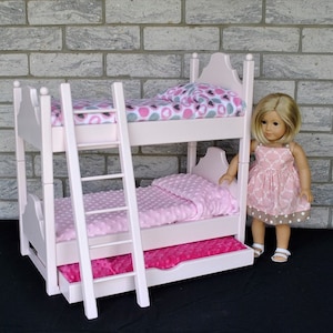 May include: A white wooden bunk bed with a pink and gray patterned blanket on the top bunk and a pink blanket on the bottom bunk. The bed has a ladder and a pull-out drawer. A doll is standing next to the bed.