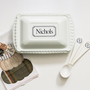 May include: White ceramic baking dish with a scalloped edge and the word "Nichols" written on the front. The dish is accompanied by three white ceramic spoons with a blue "N" on the handle.