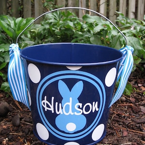 May include: Blue metal bucket with white polka dots and a blue bunny with the name "Hudson" in white lettering. The bucket has a silver handle.