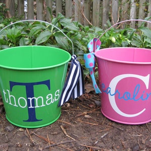 May include: Two personalized metal buckets with handles. The green bucket has a white name "thomas" with a blue "T". The pink bucket has a white name "caroline" with a white "C". Both buckets have black and white striped ribbon handles.