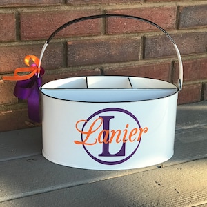 May include: White metal caddy with a black handle and a purple and orange design. The caddy has three compartments and says "Lanier" with a purple "L" in a circle.