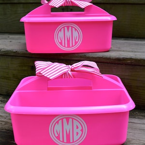 May include: Two bright pink caddies with handles and white monogrammed initials. Each caddy has a white and pink striped bow on top. The caddies are sitting on wooden steps.