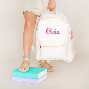 Op de afbeelding: Witte rugzak met gouden ritsen en de naam "Olivia" in roze script. Een roze potlood zit in de zijzak. De persoon draagt gouden sandalen en staat op twee boeken, één turquoise en één lavendel.