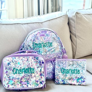 May include: A set of three school accessories: a backpack, lunch bag, and pencil case. Each item is clear with a purple border and filled with colorful glitter. The name "Charlotte" is printed in teal on each item.