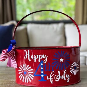 May include: A red metal bucket with a red handle featuring the white and blue text Happy 4th of July, white and blue firework graphics, and a decorative bow made of blue ribbon and red and white striped ribbon attached to the side handle.