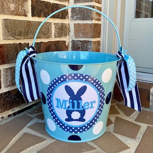 May include: A light blue metal Easter basket with a white and navy blue polka dot pattern. The basket has a white circle with a navy blue border and a navy blue bunny silhouette with the name "Miller" in white text. The basket has a white and navy blue striped ribbon attached to the handle.
