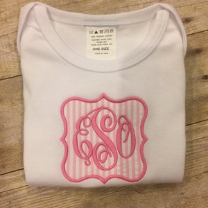 Könnte beinhalten: Ein weißer Baby-Strampler mit einem gestickten rosa Monogramm auf der Vorderseite. Das Monogramm befindet sich in einem rosa-weiß gestreiften Rahmen. Der Strampler hat ein Etikett mit der Aufschrift "100% Bio-Baumwolle, Maschinenwäsche kalt, Trocknergeeignet, Warm bügeln auf links, Einheitsgröße, Hergestellt in Indien."