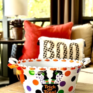 May include: White metal Halloween candy bucket with a scalloped rim and orange ribbon. The bucket is decorated with colorful polka dots and a witch's legs sticking out of a cauldron with the text "Trick or Treat".