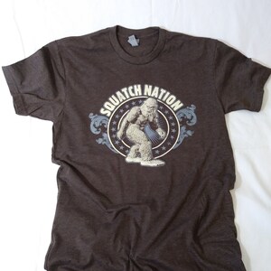 May include: A brown t-shirt with a graphic of a Bigfoot in a circle with the text "SQUATCH NATION" around it.
