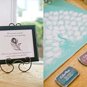 May include: A framed sign with a peacock feather illustration and text, displayed on a decorative stand. The sign reads, "We're proud as a peacock to have you part of our special day. Please leave your fingerprint and sign your name inside a feather on our guest book." A teal peacock guest book and ink pads are also visible.