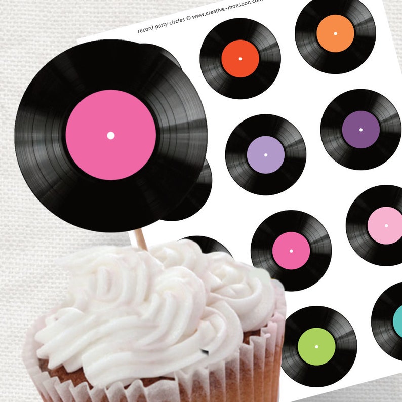 May include: A cupcake topped with white frosting and a black and white vinyl record cupcake topper. The record has a pink center.
