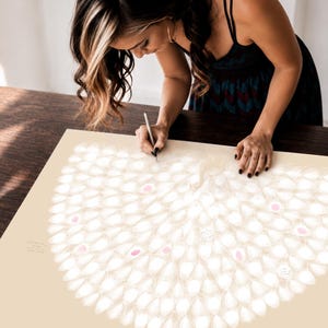 Woman signing a personalised peacock thumbprint guest book poster at a baby shower, featuring cream peacock feathers with soft pink fingerprint details and handwritten messages.