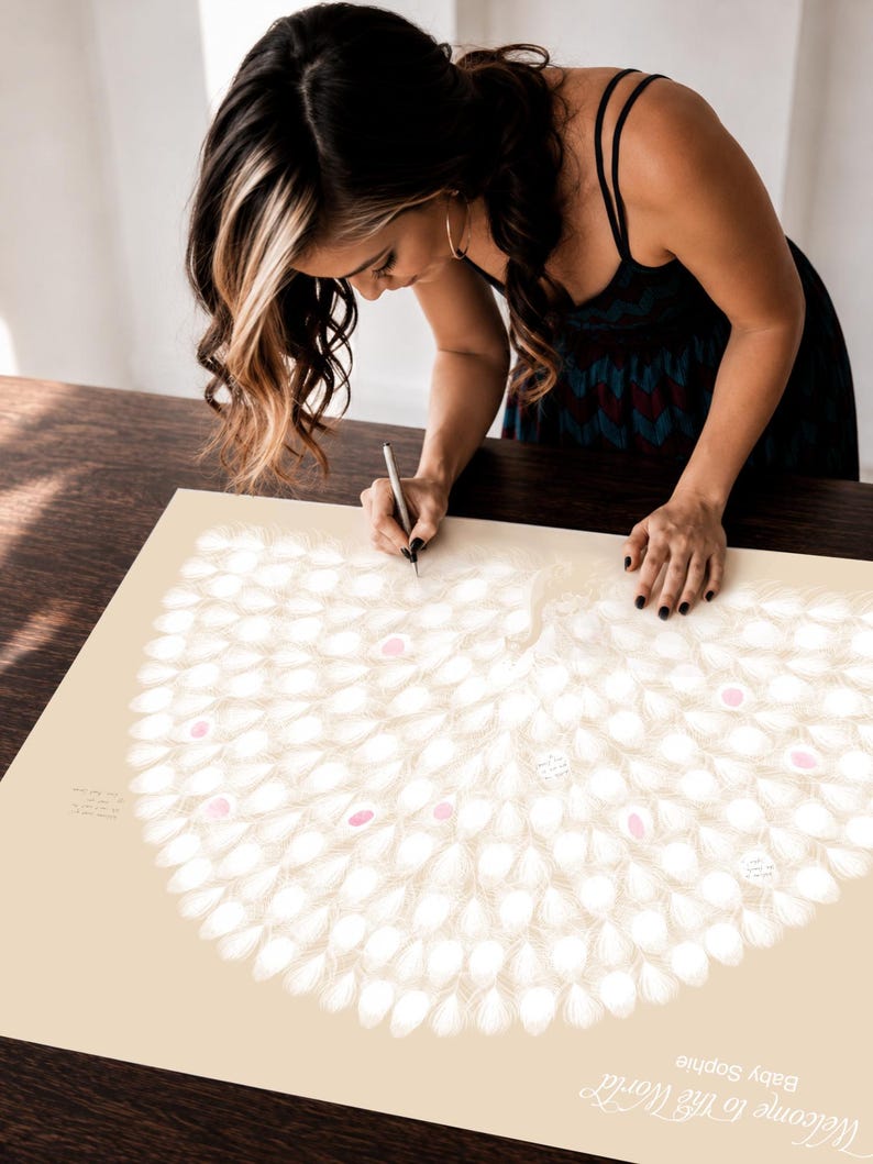 Woman signing a personalised peacock thumbprint guest book poster at a baby shower, featuring cream peacock feathers with soft pink fingerprint details and handwritten messages.