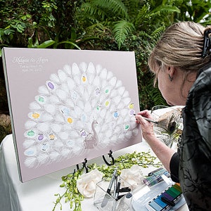 May include: A light purple wedding guest book with a peacock design. The peacock's feathers are white with colorful thumbprints. The names "Melanie & Jason" and the date "April 28, 2012" are printed at the top.