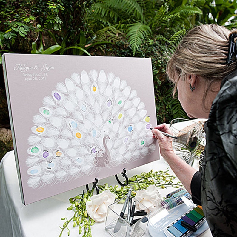 May include: A light purple wedding guest book with a peacock design. The peacock's feathers are white with colorful thumbprints. The names "Melanie & Jason" and the date "April 28, 2012" are printed at the top.