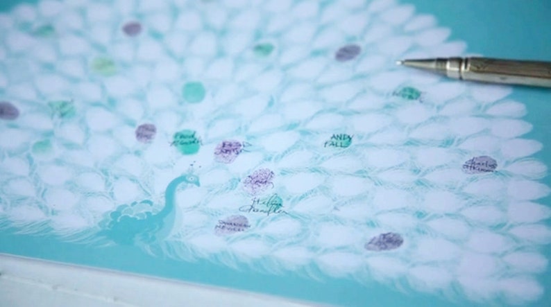 May include: A teal peacock-themed guest book with a pen. The peacock's tail feathers are represented by white shapes, with colorful fingerprints and names. The peacock is on a light blue background.