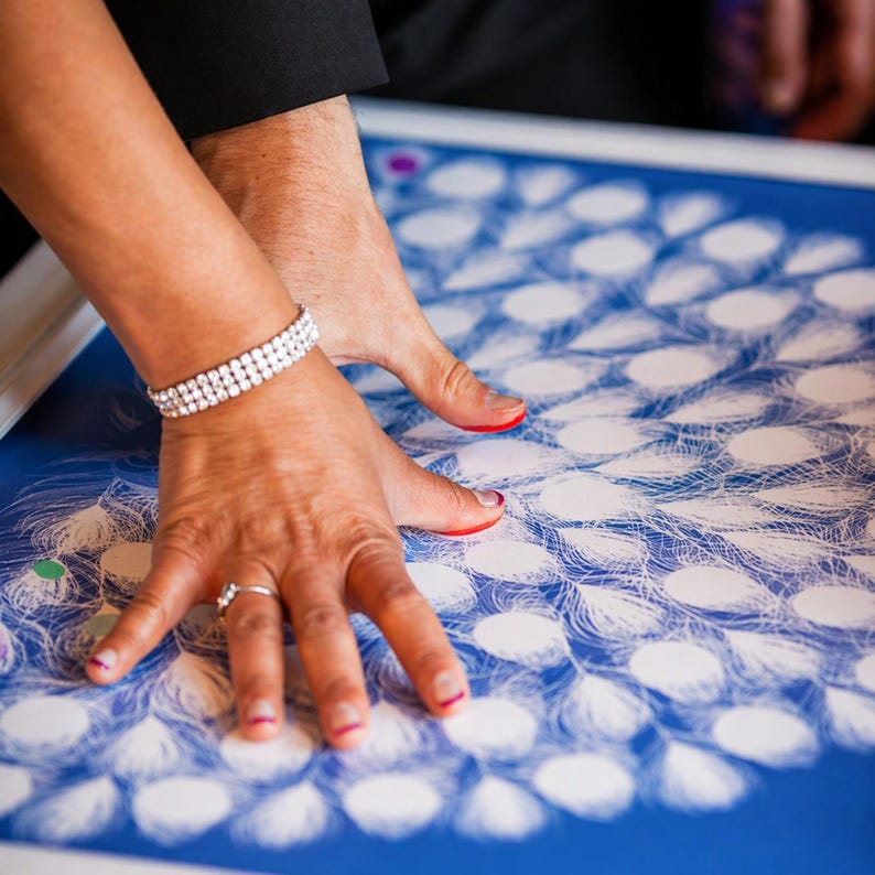 May include: A blue and white guest book alternative with a peacock feather design. Two hands are placed on the surface, with a diamond bracelet and ring visible. The design is a unique alternative to a traditional guest book.