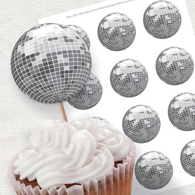 May include: A cupcake topped with white frosting and a silver disco ball cupcake topper. The cupcake topper is a printed image of a disco ball with a silver mirrored finish.