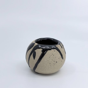 May include: A small, round ceramic vase with a speckled, light tan exterior. The vase features a black interior and is adorned with black, dripped paint-like designs. The overall aesthetic is minimalist and artistic.