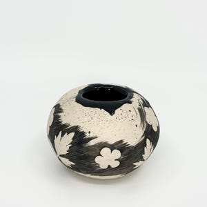 May include: A round ceramic vase with a cream-coloured base featuring black brushstroke patterns and white floral and leaf silhouettes. The rim is finished with a thick, glossy black glaze, and the surface has a speckled texture throughout.