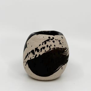 Op de afbeelding: Een keramische vaas met een afgeronde vorm. De vaas is beige met een groot zwart penseelstreekontwerp en zwarte verfspatten. De rand van de vaas is ook zwart geschilderd. De vaas heeft een moderne, artistieke uitstraling.