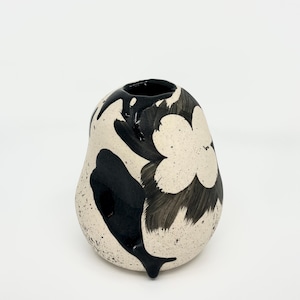 May include: A bulbous ceramic vase with a cream-coloured base featuring black speckled details. The surface is decorated with bold, hand-painted black floral motifs and thick, glossy black glaze drips extending from the circular opening downwards.
