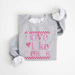 Puede incluir: Una sudadera gris con la frase "Love Like Jesus" en letras rosas. El texto incluye un coraz&oacute;n y est&aacute; enmarcado por un patr&oacute;n de cuadros rosas. Las mangas est&aacute;n enrolladas, revelando el interior blanco.