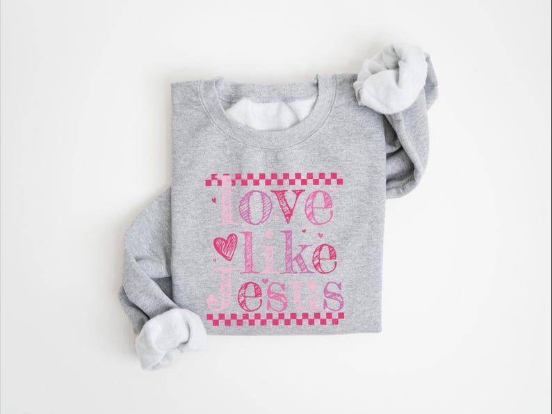 Puede incluir: Una sudadera gris con la frase "Love Like Jesus" en letras rosas. El texto incluye un coraz&oacute;n y est&aacute; enmarcado por un patr&oacute;n de cuadros rosas. Las mangas est&aacute;n enrolladas, revelando el interior blanco.