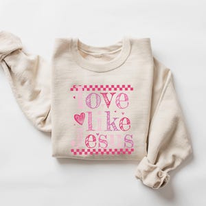 Puede incluir: Sudadera beige con las palabras "Love like Jesus" en letras rosas y moradas. La palabra "love" est&aacute; encima de un coraz&oacute;n, y el texto est&aacute; enmarcado por patrones de cuadros rosas. Mangas enrolladas.