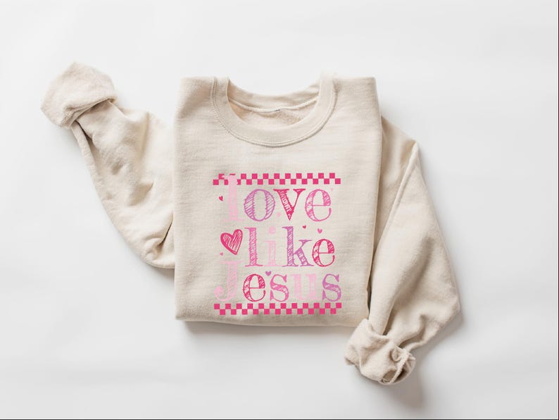 Puede incluir: Sudadera beige con las palabras "Love like Jesus" en letras rosas y moradas. La palabra "love" est&aacute; encima de un coraz&oacute;n, y el texto est&aacute; enmarcado por patrones de cuadros rosas. Mangas enrolladas.