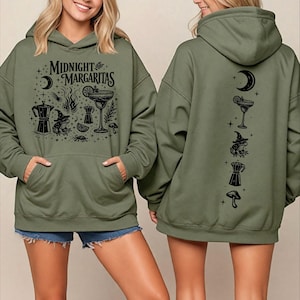 May include: An olive green hooded sweatshirt featuring a black graphic print. The front displays the text "MIDNIGHT MARGARITAS" with illustrations of a margarita glass, a frog in a witch hat, a moka pot, a crescent moon, and mushrooms.
