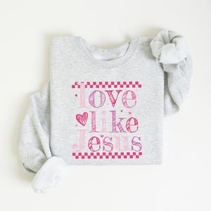 Puede incluir: Sudadera gris claro con la frase "Love like Jesus" en rosa. El texto est&aacute; enmarcado por un borde rosa a cuadros. La sudadera est&aacute; doblada, con las mangas enrolladas.