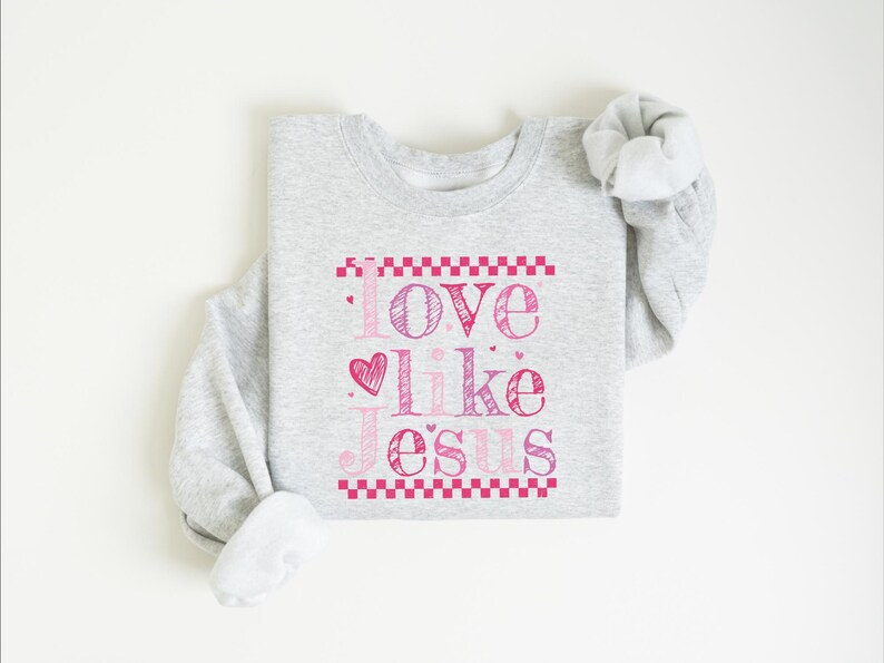 Puede incluir: Sudadera gris claro con la frase "Love like Jesus" en rosa. El texto est&aacute; enmarcado por un borde rosa a cuadros. La sudadera est&aacute; doblada, con las mangas enrolladas.
