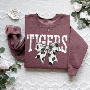 A maroon sweatshirt with a white and black polka dot bow on it, placed on a white surface with some flowers and a small white object nearby.