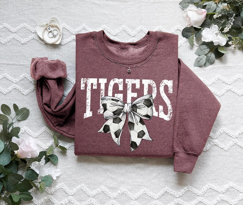 A maroon sweatshirt with a white and black polka dot bow on it, placed on a white surface with some flowers and a small white object nearby.