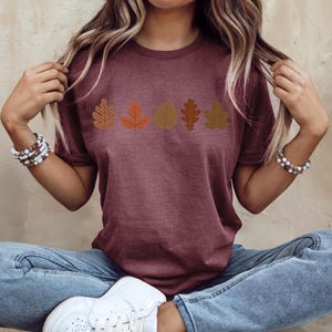 A woman wearing a maroon t-shirt with a graphic of autumn leaves, sitting cross-legged with her hands behind her head.